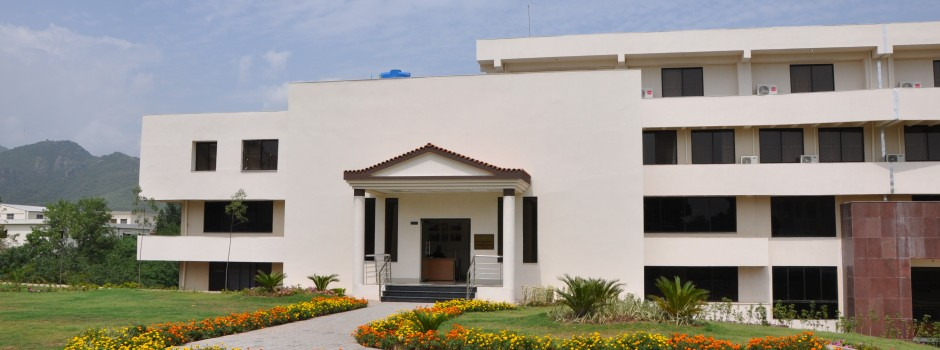 Front view of Bahria University Hostel building, featuring a neatly landscaped lawn with flowers and a backdrop of distant mountains. The building is three stories high with a main entrance that has a small porch and stairs leading to the door.