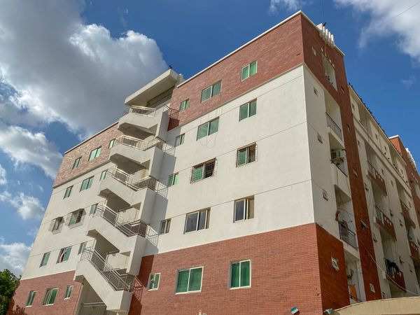 A multi-story Iqra University hostel building with red brick and white facade, featuring several windows and an external staircase, under a partly cloudy blue sky.