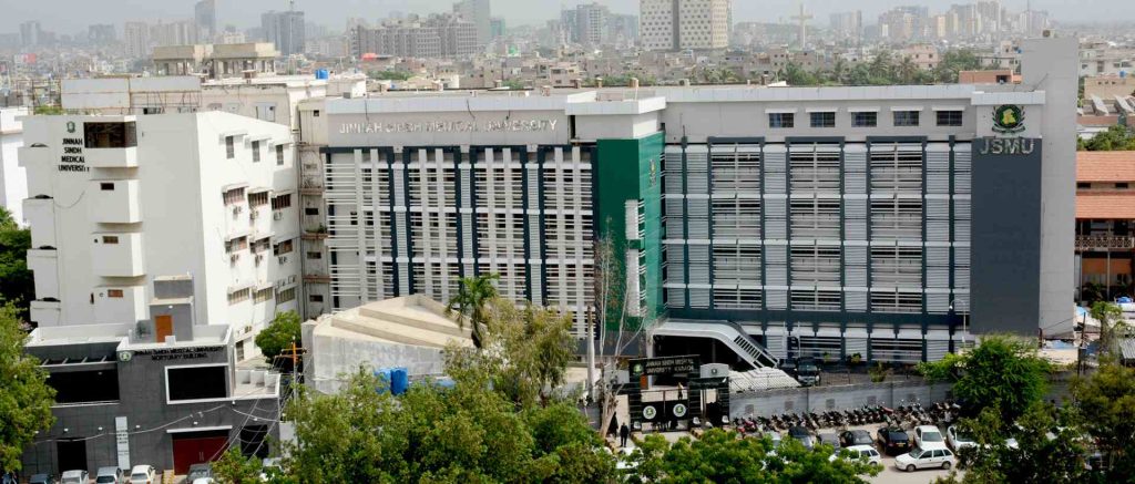 An aerial view of Jinnah Sindh Medical University (JSMU) in Karachi, featuring a large modern building with a green accent facade, surrounded by greenery and cityscape in the background, with the university's name prominently displayed on the structure