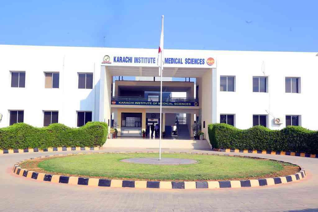 A bright and clean facade of the Karachi Institute of Medical Sciences (KIMS) building, featuring a two-story entrance with the institute's name displayed above, surrounded by neatly trimmed hedges and a central flagpole in front.